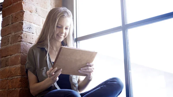 Jovem sorrindo mulher atraente feliz segurando um tablet digital sentado no peitoril da janela — Fotografia de Stock