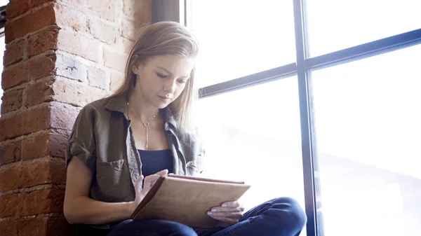 Young woman working with a digital tablet while sitting on the window sill — Stock Photo, Image