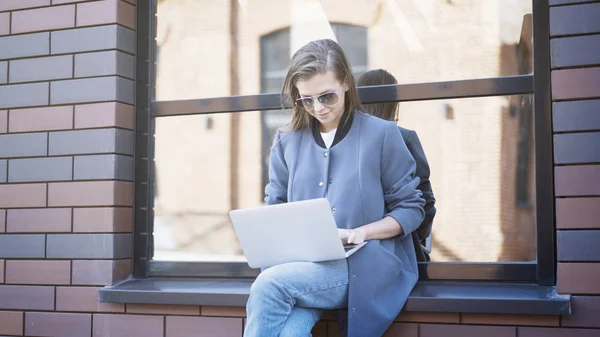 Una giovane ragazza sorridente in occhiali da sole è seduta su un davanzale a digitare in un computer portatile — Foto Stock