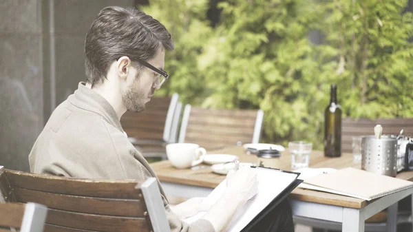 Un hombre europeo guapo con gafas haciendo notas dibujando en la cafetería al aire libre — Foto de Stock