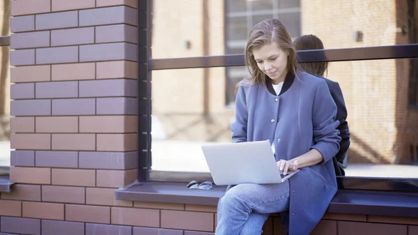 Una giovane ragazza sorridente è seduta su un davanzale a digitare un computer portatile — Foto Stock