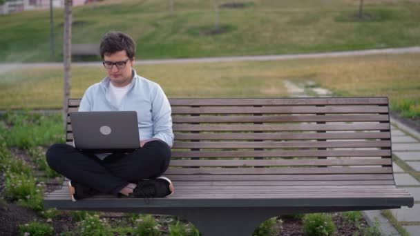 Mladý muž pomocí notebooku v parku sedí na lavičce na rozmazané trávě pozadí — Stock video