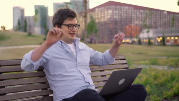 Jovem termina o trabalho em laptop sentado em um banco no fundo do parque desfocado — Vídeo de Stock