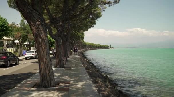 Gardasee - 18. Juli: Echtzeit-Weitschuss vom Ufer des Gardasees. Menschen gehen entlang der Böschung und bewundern die Landschaft, den Gardasee. — Stockvideo