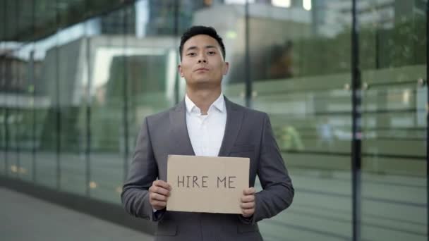 Left to right pan real time shot of a young Asian man in a suit standing on the street with a sign "hire me". — Stock Video