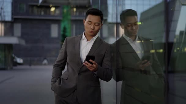 Real time shot of a young asian businessman conducting business correspondence, standing leaning against the wall. — 비디오