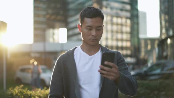 Real time shot of a young asian man standing on the street and leading a correspondence on the phone. — Stock video