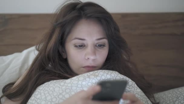 손으로 찍은 여자가 침대에 누운 채 전화기를 검색하는 모습 — 비디오