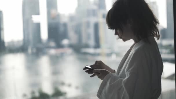 Gelişmiş bir otelin panoramik penceresinin önündeki akıllı telefonu kullanan bir kadın. Arka planda Singapur silueti. gerçek zamanlı çekim. — Stok video