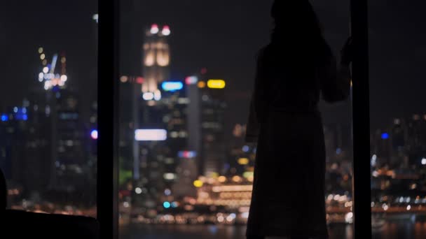 Fechado tiro de uma mulher de negócios em roupão de banho olhando para o horizonte moderno em Singapura à noite. Imagens em tempo real da paisagem urbana do centro em segundo plano — Vídeo de Stock