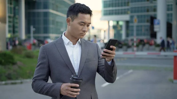 Портрет молодого азіата-бізнесмена, що веде кореспонденцію по телефону з кавою в руках. Центральний район. — стокове фото