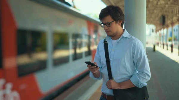 Молодий бізнесмен у блакитній сорочці та окулярах пише, стоячи на платформі, чекаючи на свій потяг. Сонячний літній день . — стокове фото