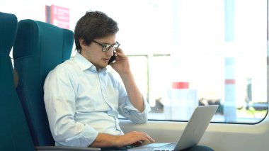 Beyaz gömlek ve kot pantolon giyen genç müdür işe trenle gidip bilgisayarını kullanıyor..