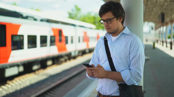 Молодий бізнесмен у блакитній сорочці та окулярах пише, стоячи на платформі, чекаючи на свій потяг. Сонячний літній день . — стокове фото