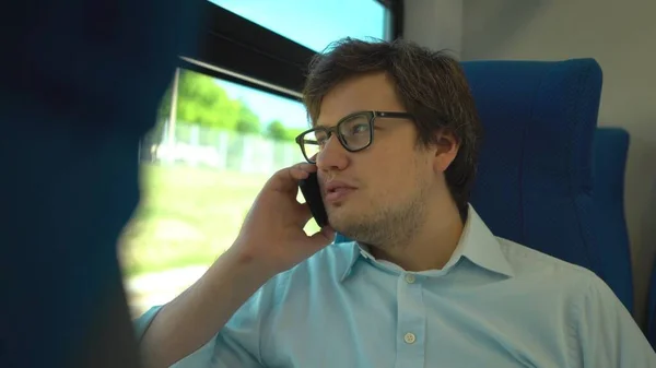 Bonito jovem empresário vestindo uma camisa azul e óculos está montando um trem discando um número e falando em seu telefone. Dia de verão ensolarado . — Fotografia de Stock