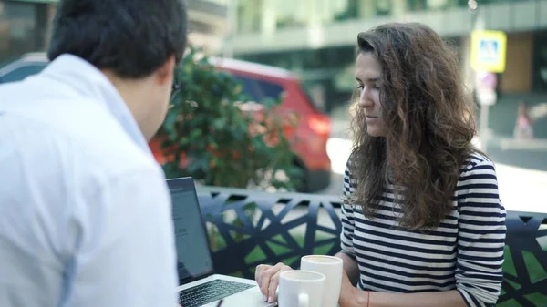 Mulher nova na camisa listrada e seu colega bonito na camisa branca que trabalha junto no parque do verão usando o portátil e a prancheta. O homem está a beber café. . — Fotografia de Stock