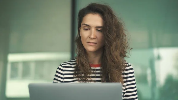 Attraente giovane donna con lunghi capelli ricci che indossa camicia a righe è seduto sulle scale di costruzione in città e laptop di lavoro — Foto Stock