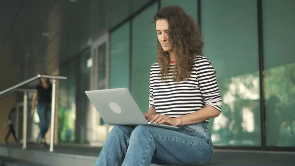 Uzun kıvırcık saçlı, çizgili tişörtlü çekici bir kadın şehirdeki bir binanın merdivenlerinde oturuyor ve dizüstü bilgisayarla çalışıyor. — Stok fotoğraf