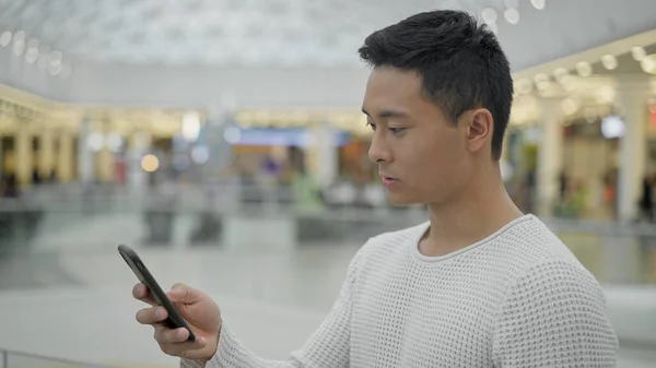 Портрет азіатського чоловіка, що прокручує телефон на фоні великого торгового центру — стокове фото