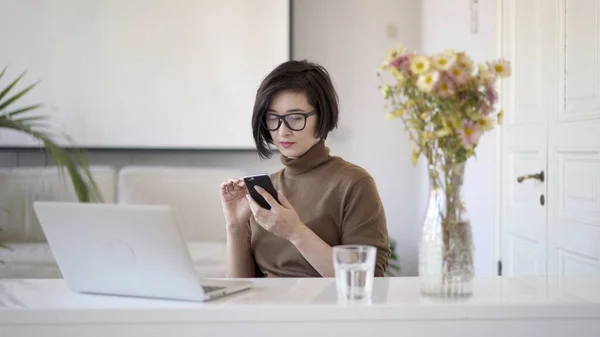Aziatische vrouw in bril werken met laptop in wit home office en surfen internet in smartphone — Stockfoto