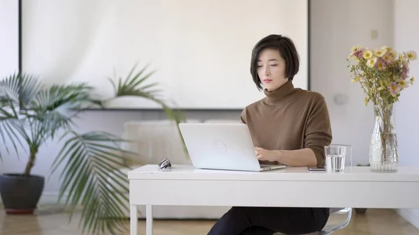 Asiatisk kvinne i briller som arbeider med bærbar PC på hvit hjemmekontor – stockfoto