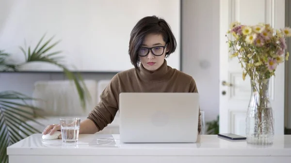 Asiatisk kvinne i briller som arbeider med bærbar PC på hvit hjemmekontor – stockfoto