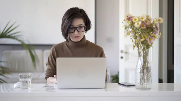 Asiatisk kvinne i briller som arbeider med bærbar PC på hvit hjemmekontor – stockfoto