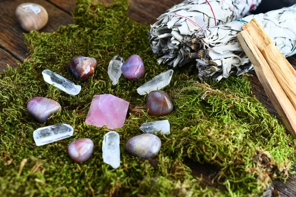 A close up image of a crystal energy healing grid on moss with various smudge sticks.