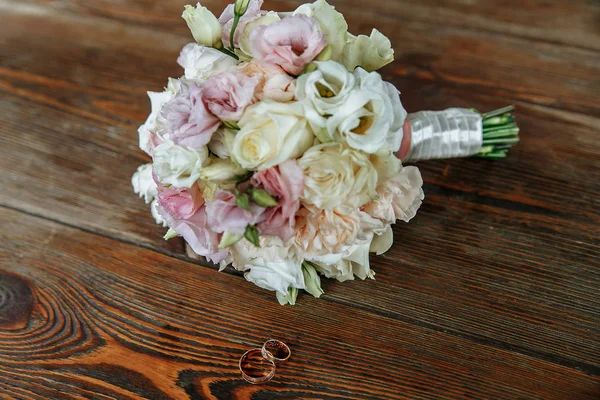 Hochzeitsstrauß aus Creme und rosa Rosen liegt auf einer Holzoberfläche. Eheringe — Stockfoto
