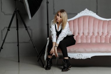 Beyaz gömlekli ve siyah tulumlu güzel bir kız. Pembe bir kanepede. Moda fotoğrafçılığı üzerine bir fotoğraf çekiminde.