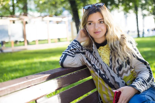 Technology smartphone nature outdoor concept. Girl listening to music. Young lady in park sitting on bench enjoying sound wearing headphones.