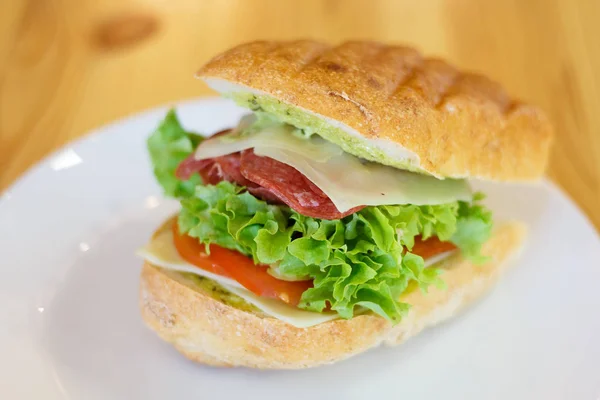 Sandwich with salami, cheese, basil and pesto sauce on a white plate. Beautifully served sandwich on a wooden table. Selective focus — Stock Photo, Image