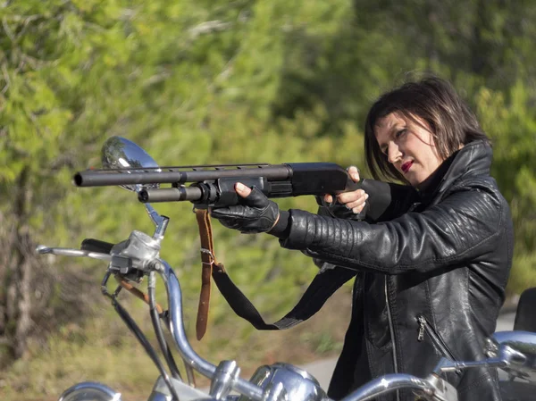 A woman in a black leather biker jacket with a carbine rifle on a chopper motorcycle in Greece on a road in the forest in the mountains