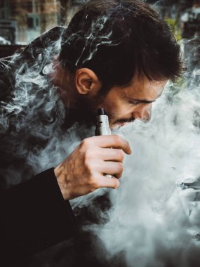 Bir adam elini tutuyor ve elektronik bir sigara içiyor. VAPE ve buhar bulutları salıyor.)