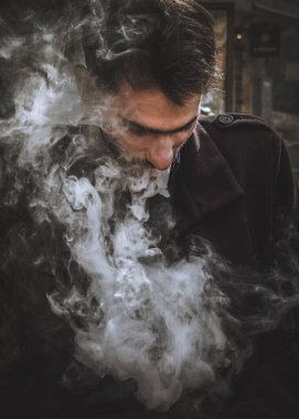 Bir adam elini tutuyor ve elektronik bir sigara içiyor. VAPE ve buhar bulutları salıyor.)