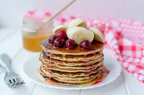 Desayuno saludable. Pila de deliciosos panqueques caseros con miel, cereza y plátano en plato blanco — Foto de Stock