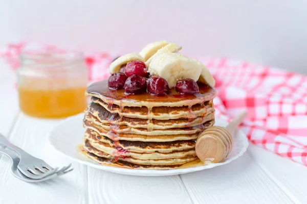 Desayuno saludable. Pila de deliciosos panqueques caseros con miel, cereza y plátano en plato blanco — Foto de Stock