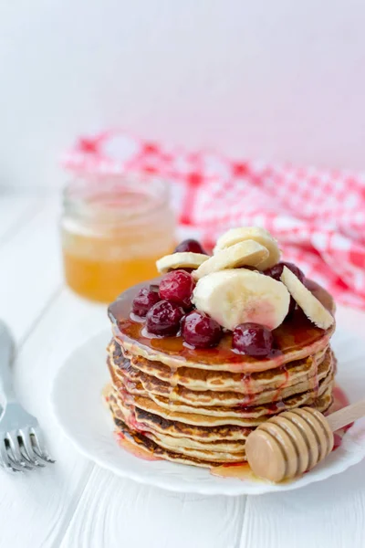 Desayuno saludable. Pila de deliciosos panqueques caseros con miel, cereza y plátano en plato blanco — Foto de Stock
