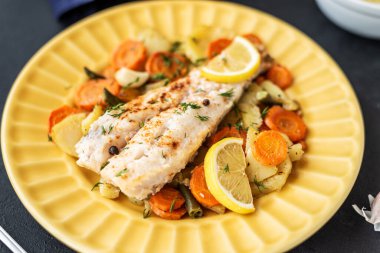 Fırında pişmiş hake balığı ve siyah arka planda sarı bir tabakta sebzeler. Yemekler için lezzetli sıcak atıştırmalıklar. Seçici odak