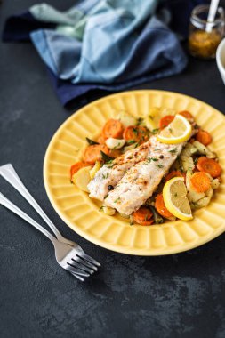 Fırında pişmiş hake balığı ve siyah arka planda sarı bir tabakta sebzeler. Yemekler için lezzetli sıcak atıştırmalıklar. Seçici odak