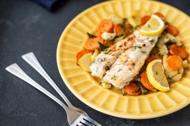 Fırında pişmiş hake balığı ve siyah arka planda sarı bir tabakta sebzeler. Yemekler için lezzetli sıcak atıştırmalıklar. Seçici odak