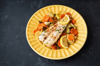 Fırında pişmiş hake balığı ve siyah arka planda sarı bir tabakta sebzeler. Yemekler için lezzetli sıcak atıştırmalıklar. Seçici odak