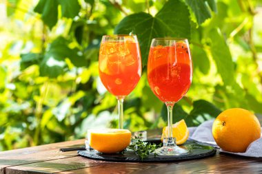Portakallı büyük şarap kadehinde iki Aperol spreyi kokteyli, İtalyan taze soğuk içeceği. Üzüm bağı olan güneşli bir bahçe.