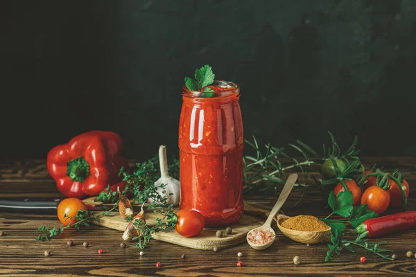 Homemade DIY natural canned hot tomato sauce chutney with chilli or adjika in glass jar standing on wooden table with cherry tomatoes, salt, pepper, herbs, selective focus — Stock Photo, Image