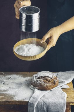 Ahşap bir masanın üzerinde un elemekte olan bir kadın. Koyu renk önlüklü şef, ekmeği koyu bir arka planda hazırlamak için unu süzgeçten geçirir..
