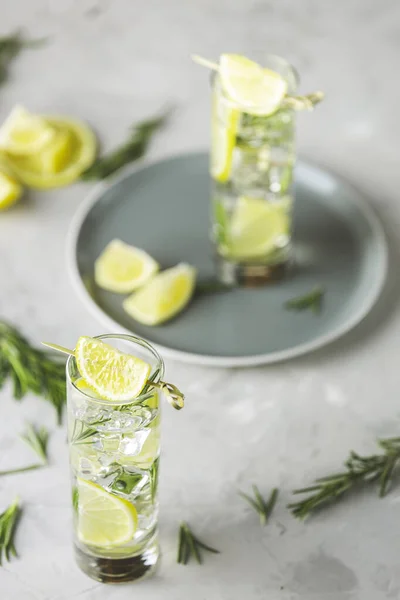 Erfrischendes Limettengetränk mit Eiswürfeln in Glasbechern vor hellgrauem Hintergrund. Sommerfrischer Limonaden-Cocktail mit Rosmarin, selektiver Fokus. — Stockfoto