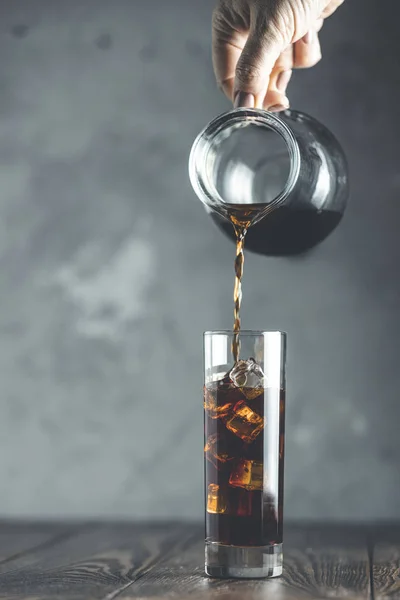 Frauenhand gießt selbstgebrühten kalten Kaffee aus einem kleinen Glas in ein Glas mit Eis. kaltes Sommergetränk auf einem dunklen Holztisch und grauem Hintergrund mit Kopierraum. — Stockfoto