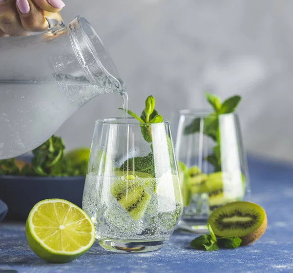 Women hand is pouring detox water, mojito or martini tonic cocktail from glass jar to glass with kiwi, lime, ice and mint. Summer fresh lime soda cocktail, selective focus.