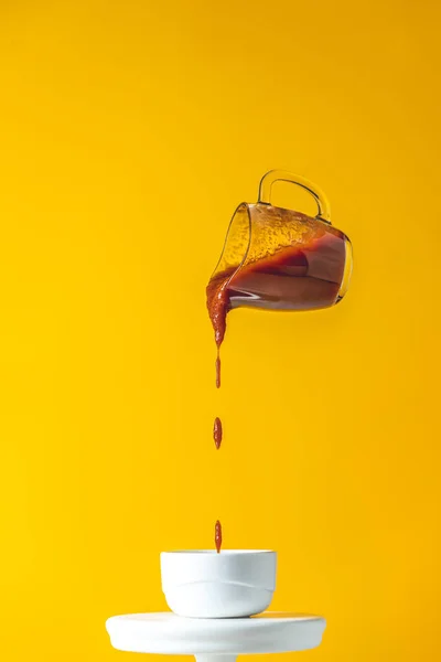 Sauce in glass jar frozen in the air. Pouring homemade DIY natural canned hot tomato sauce from small jar to white ceramic pot. Yellow food art background.