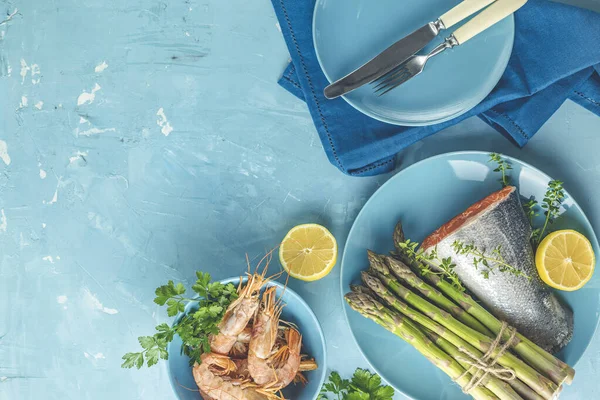 Fresh raw asparagus with trout fish, lemon and parsley, shrimp, prawn  in blue ceramic plates with napkin on light blue concrete table surface. Healthy food background with copy space for you text.